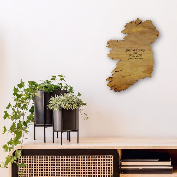 This Claddagh Ring wall art is a wooden map of Ireland. It is engraved with the Bride and Groom's names, the date of their wedding, and a Celtic claddagh ring symbol. The Ireland map hanging is on a wall beside two black modern plant posts that are sitting on a wooden desk.