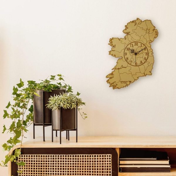 An engraved map of Ireland clock made from wood and hung on a wall. Modern black plant pots with ivy sit below it on a wooden desk.