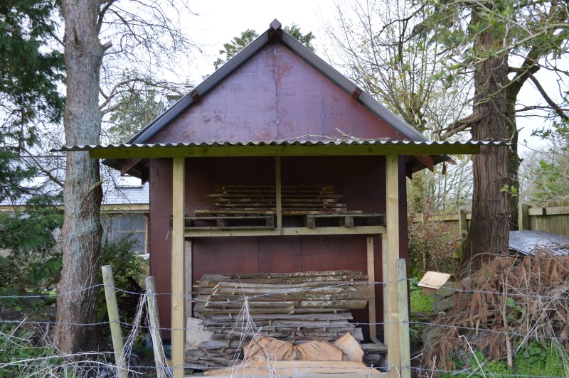 Wooden planks air-drying under a tin roof lean-to of the back of an A-frame wooden cabin