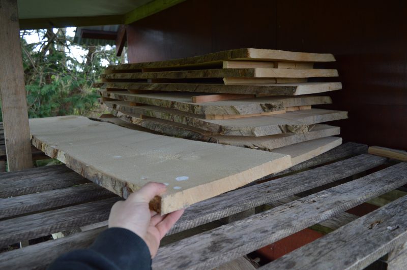 A hand is selecting a plank of timber from a stack of wooden planks that are air-drying oustide under a tin-roofed lean-to.