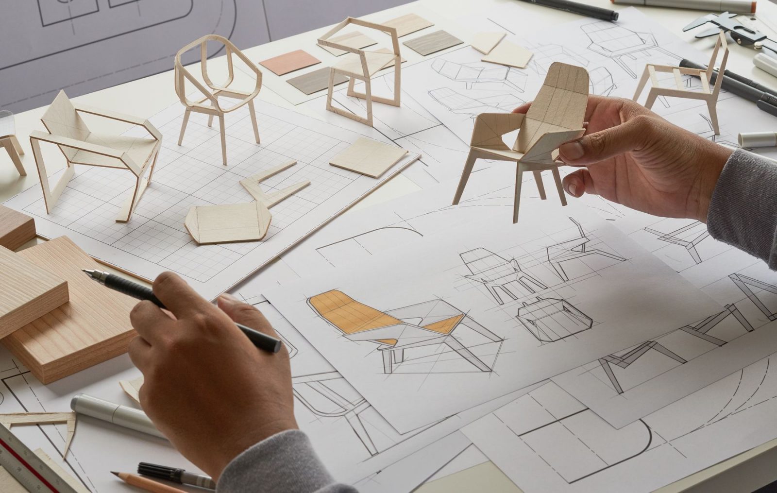 Furniture designer hands holding a pencil and a miniature chair 3d model with chair design sketches in background