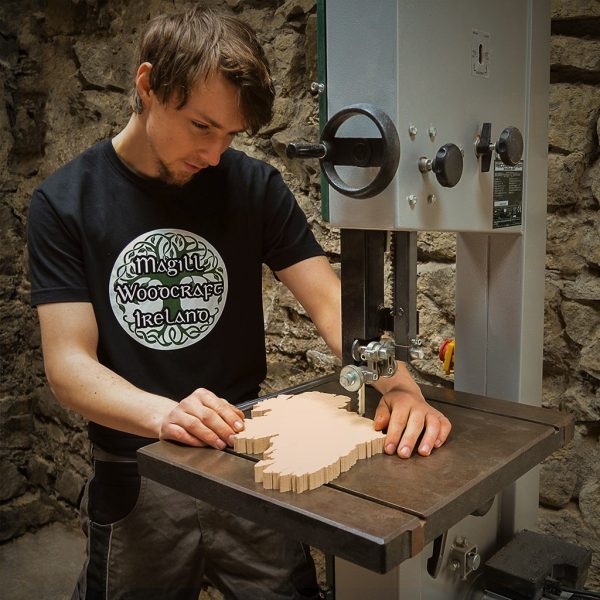 Furniture Designer-Maker Cian Magill carving a Wooden Map of Ireland Wall Art piece on Record-Power bandsaw. Cian has two hands on the wooden map and is wearing a black Magill Woodcraft Ireland t-shirt. There is a stone wall in the background.