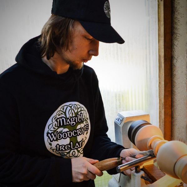 Furniture Designer Maker Cian Magill making a wooden Irish wedding goblet on a Jet woodturning lathe. He is carving the goblet to shape with a chisel, and wearing a black sweatshirt and hat with a printed Magill Woodcraft Ireland logo on.
