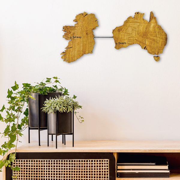 Wooden map of Ireland and Australia connected by a brass rod. The wall art piece is mounted on a white wall and above a modern/ retro wooden desk. There are also two black round modern blank pots with green hanging plants on the desk.