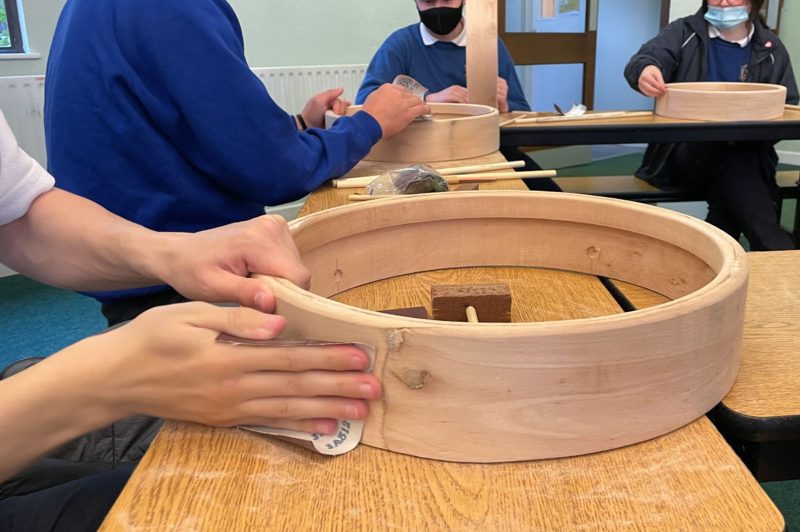 Making an Irish wooden bodhrán by hand at school in Coláiste an Eachréidh, Athenry, County Galway. Hands are sanding the bodhrán as it sits on a school desk. Other students are in the background.