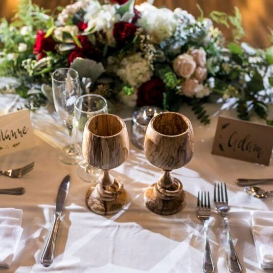 Wooden wedding goblets on white table cloth with flowers in background