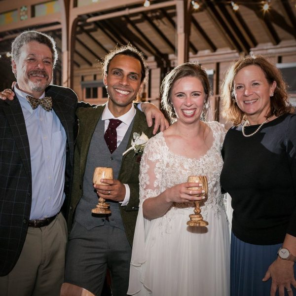Bride and groom at their wedding with parents, smilling at camera and holding two Magill Woodcraft Ireland traditonal Irish wedding goblets made from wood, with two captive rings around their stem. Interior wooden barn beams are in the background.