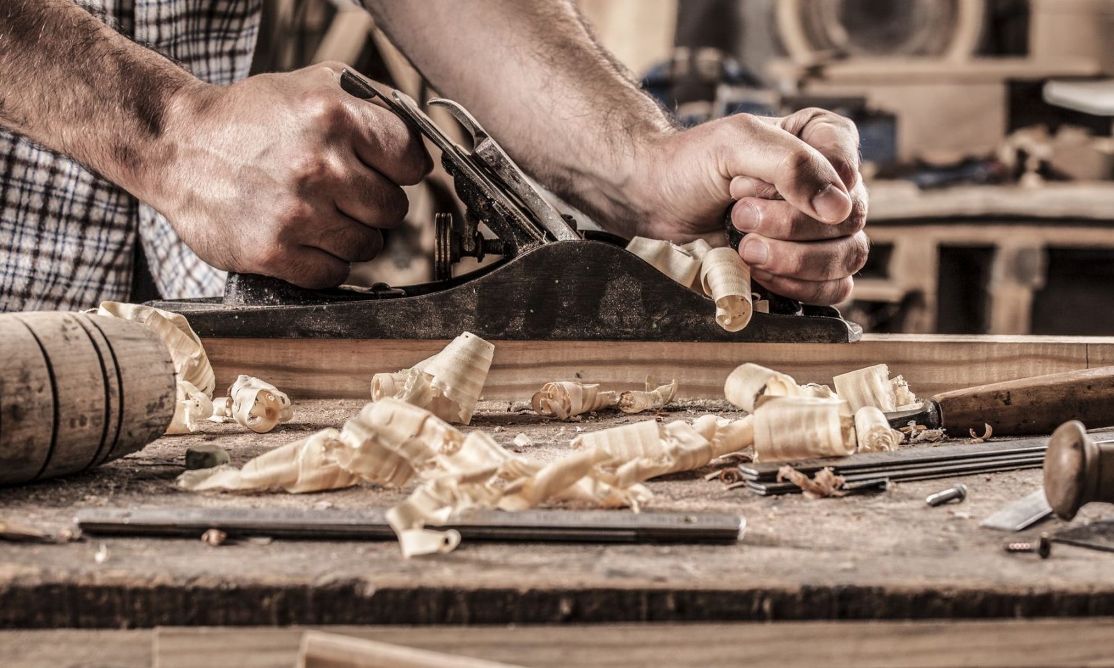 Two hands pushing a plane on wood with wood shavings and carpenter tools on workbench